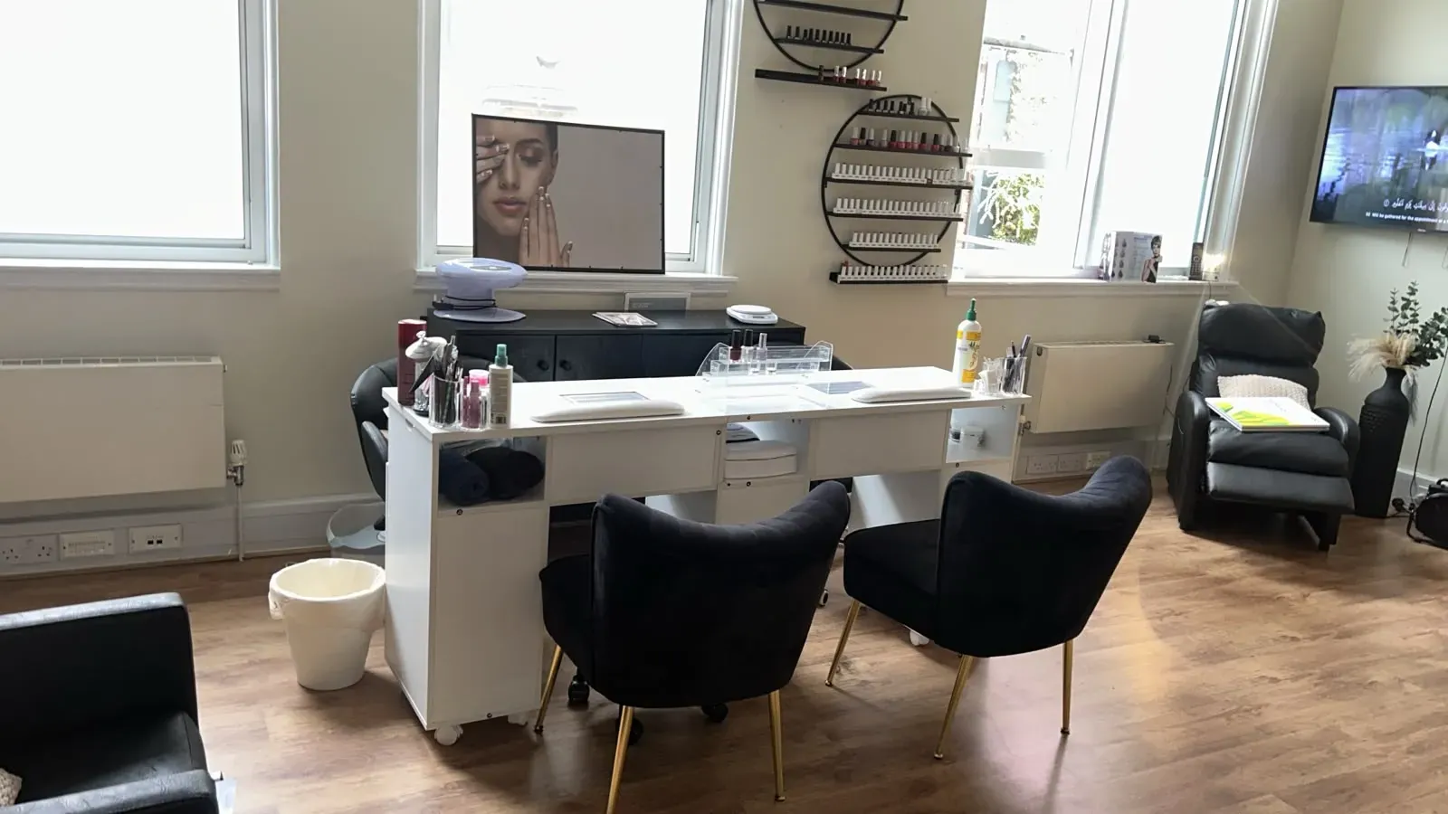 White nail desk with two velvet chairs in front of a window and a wall of nail polish